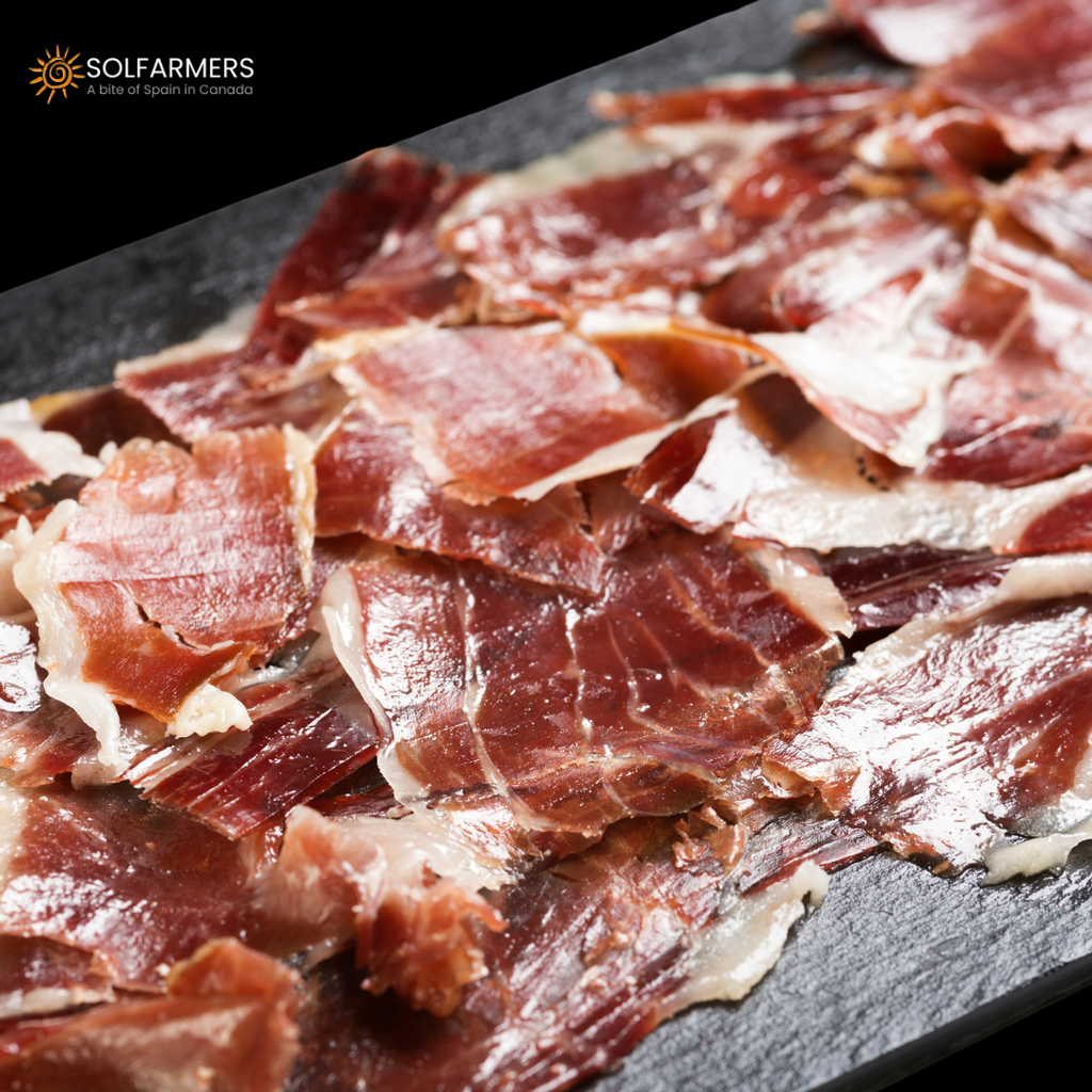 Elegant slices of Jamón Ibérico, beautifully arranged on a white plate with a touch of fresh rosemary. The rich, marbled ham highlights its melt-in-your-mouth texture and deep, savory flavors, perfect for a refined appetizer or gourmet spread