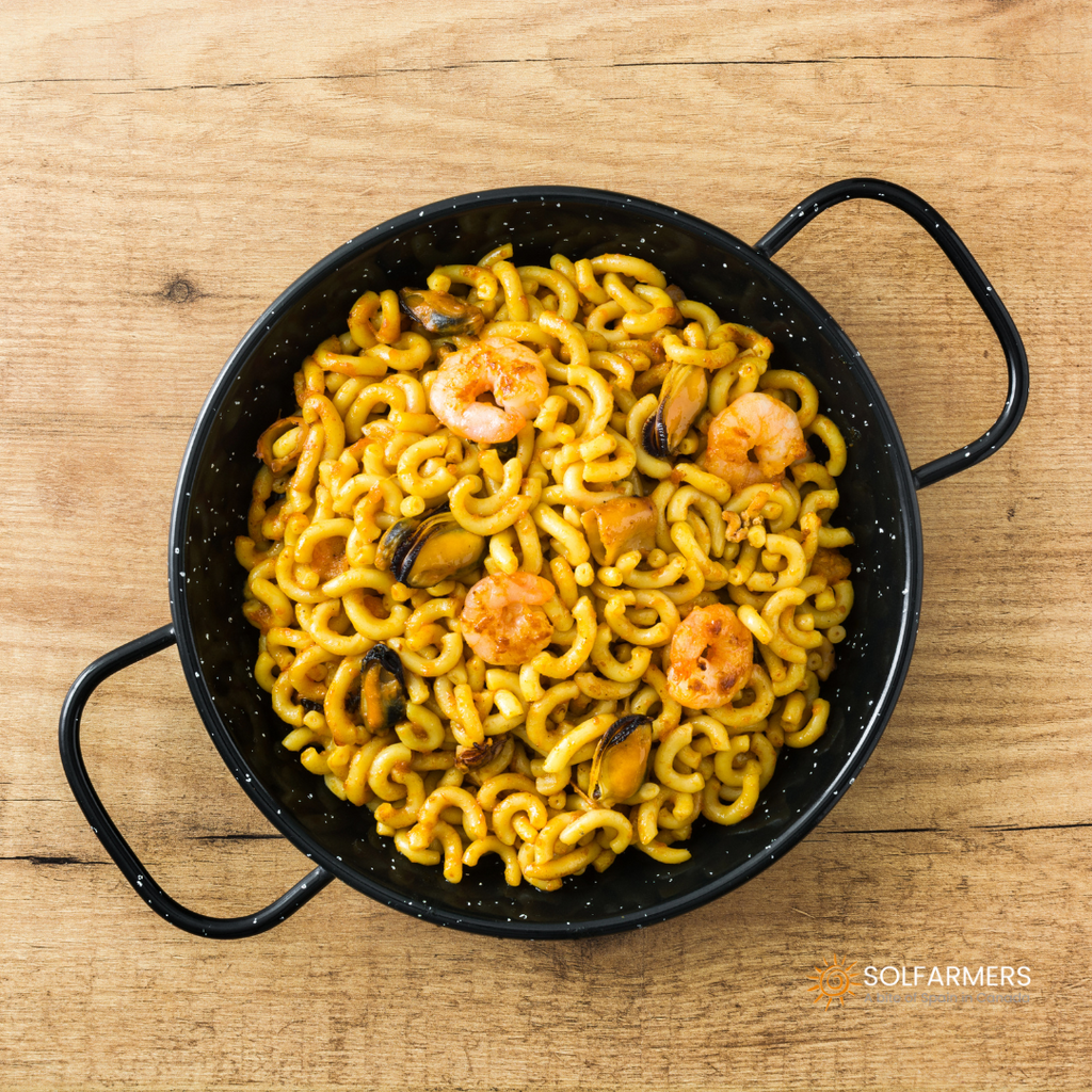 Fideuá en paellera: thin noodles browned with seafood, squid, and vegetables, cooked in a large paella pan, garnished with lemon slices and fresh parsley.