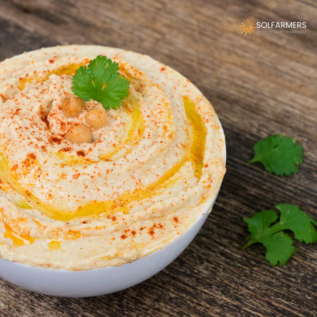 Hummus casero cremoso y suave, servido en un tazón blanco y adornado con un chorrito de aceite de oliva, pimentón, y perejil fresco picado. Perfecto como aperitivo o acompañamiento