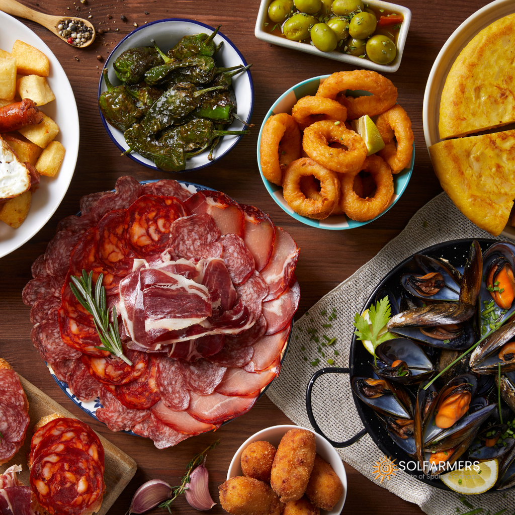  Table full of Spanish food dishes, with a variety of tapas, such as preserves, Spanish omelette, croquettes, Iberian ham, and sausages.